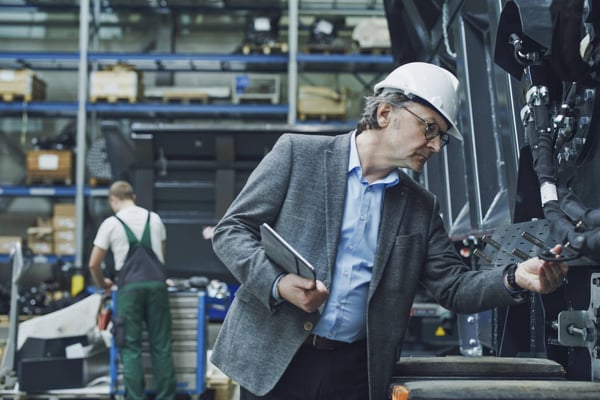 Older male employee in white hard hat assessing the efficiency of a factory machine